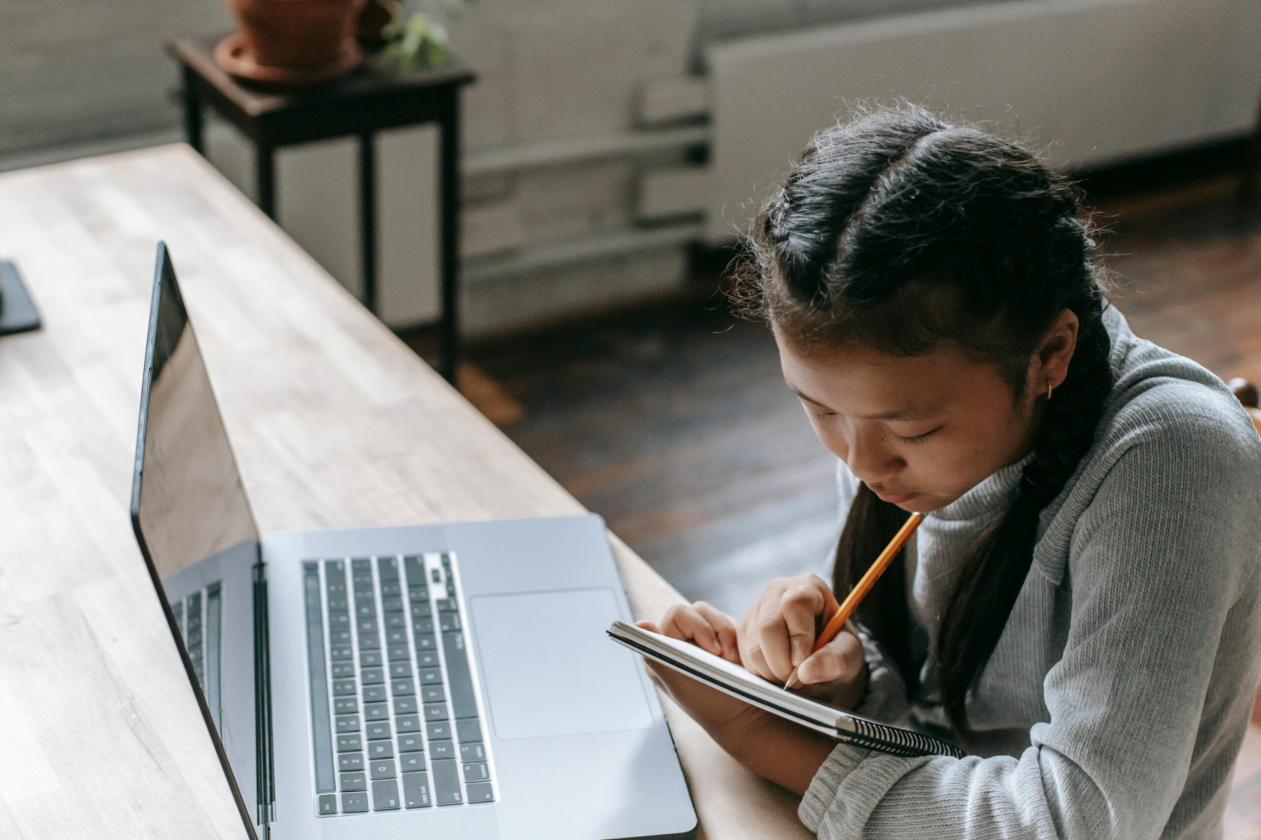 Young girl concentrating on homework with laptop, notebook, and pencil indoors.